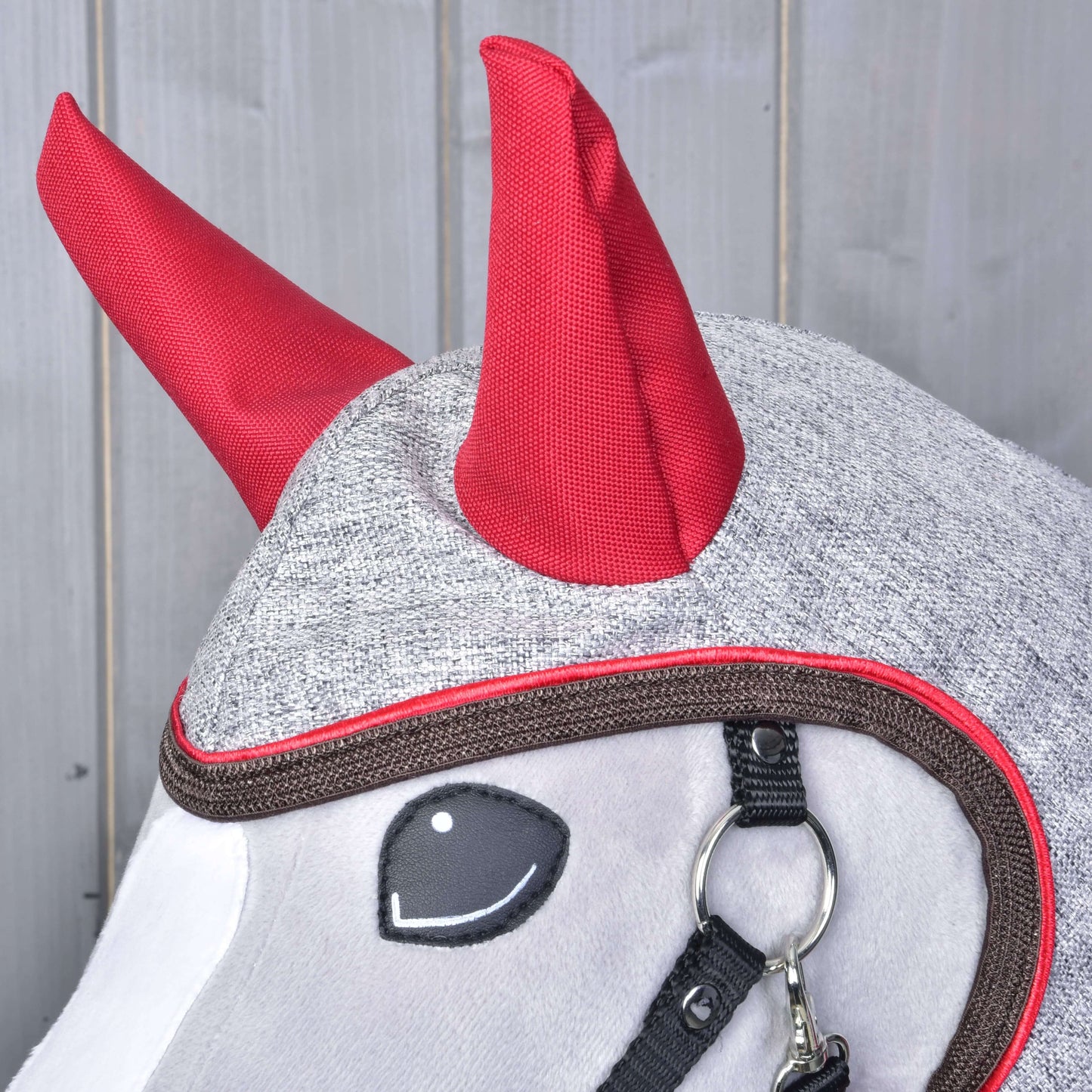 A Hobby Horse LarDen Raincoat Leonardo in grey fabric, with bright red horn-like details and black tack, is shown on a horse against a wooden background.