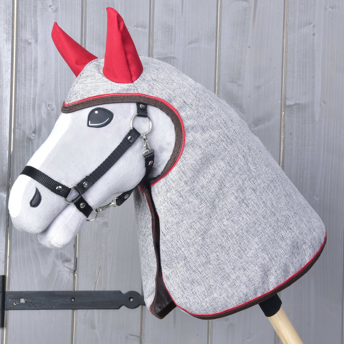 Raincoat Leonardo by Hobby Horse LarDen is a plush hobby horse with a gray head cover, red ears, and black bridle on a wooden stick—perfect for all-weather play—shown against a gray wooden wall.