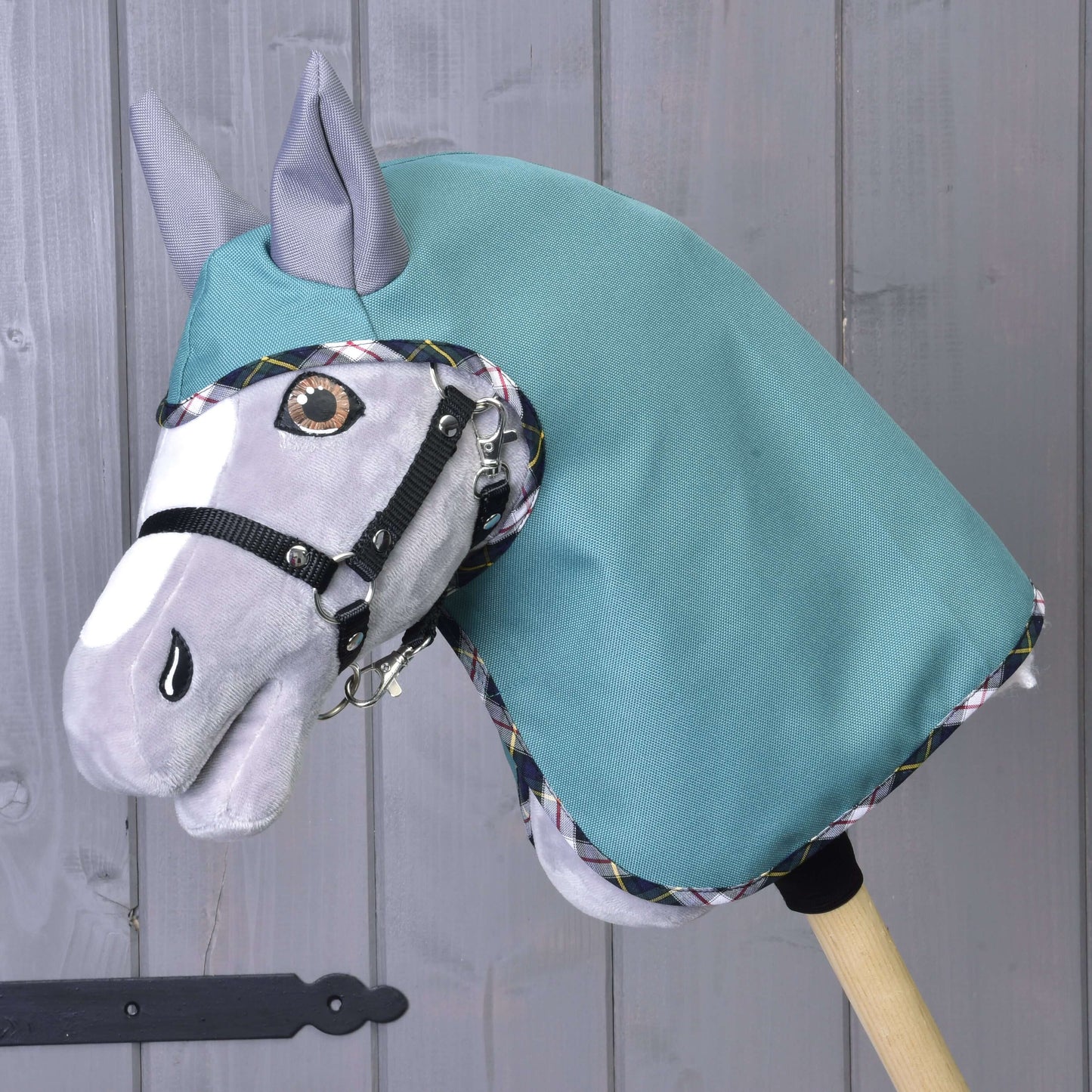 The Hobby Horse LarDen "Summer raincoat Emerald" is shown on a wooden stick, featuring a gray face, black bridle, and emerald covering in front of a gray wooden wall.