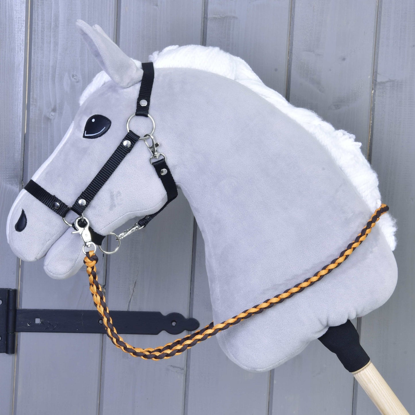The Hobby Horse LarDen gray hobby horse features a white mane, black bridle, and Reins Knitted Pomello—braided orange and brown polypropylene rope reins—mounted on a wooden stick against a gray wooden background.