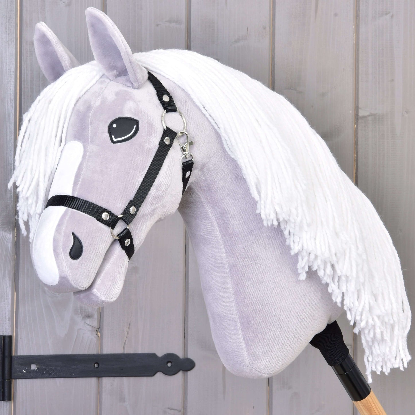 The Hobby Horse LarDen Hobby Horse Popcorn with Black Halter features a light gray head, white yarn mane, black halter, and wooden stick handle. Ideal for imaginative play and family horse adventures. Displayed against a wooden wall background.