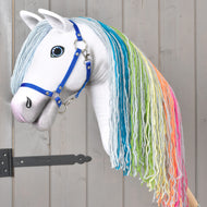 Hobby Horse Rainbow New od Hobby Horse LarDen ma białe ciało, długą tęczową grzywę z włóczki i niebieski kantar, na tle szarej drewnianej ściany z czarnym metalowym zawiasem po lewej stronie