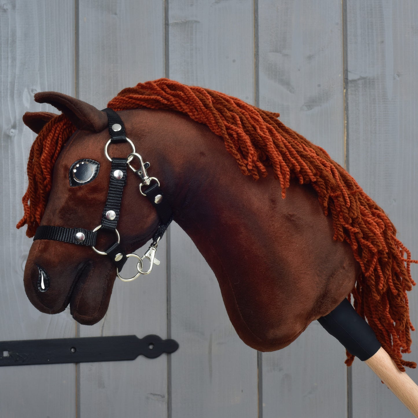 A Hobby Horse LarDen "Rebel" hobby horse in size XS, featuring a brown Hanoverian design with a yarn mane, black halter, and shiny eyes, is held in front of a gray wooden fence.
