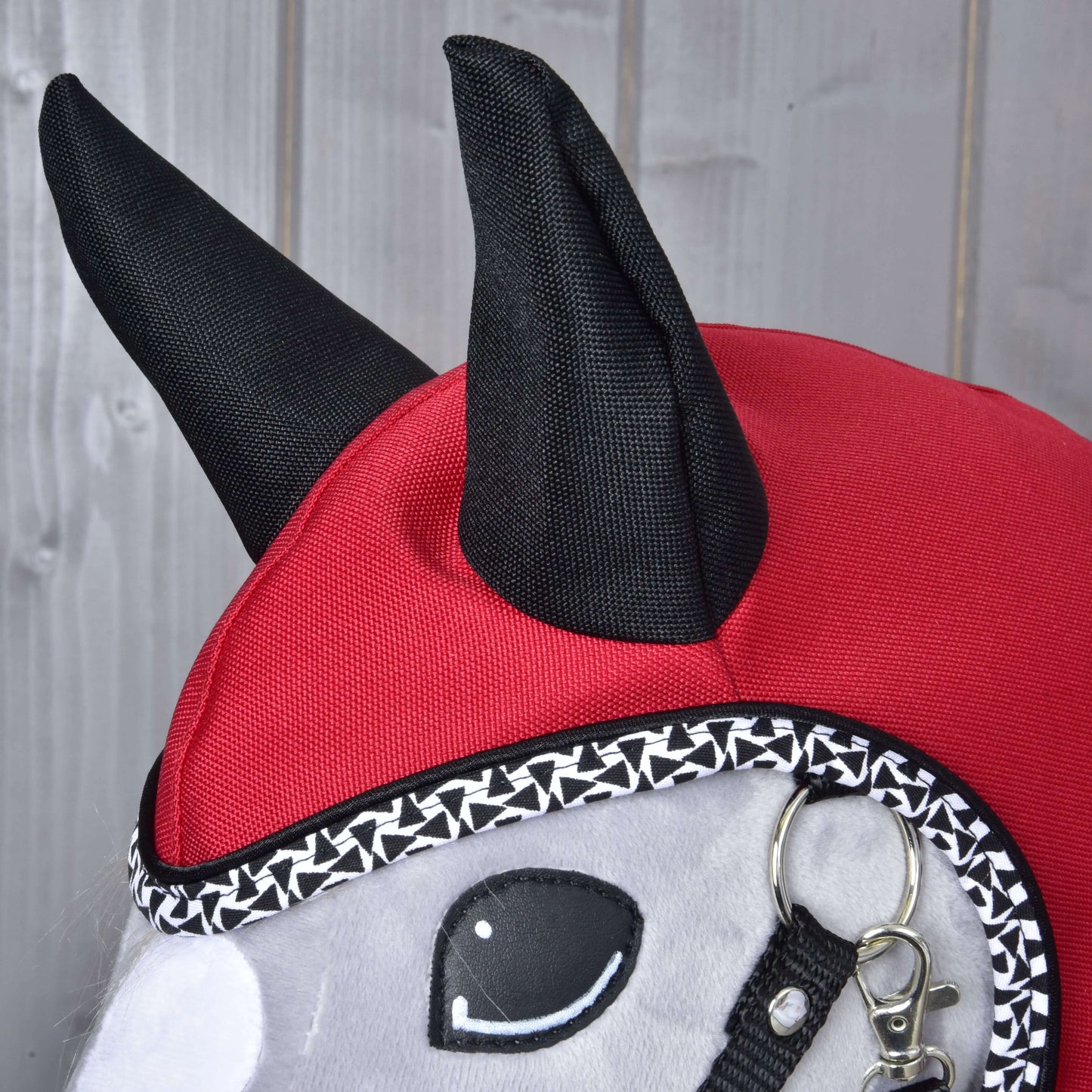 Close-up of the Hobby Horse LarDen "Raincoat Red Ferrari" plush toy, featuring a red and black hood with pointed ears, decorative trim, bridle, and stitched eyes, resembling an all-weather raincoat against a wooden background.