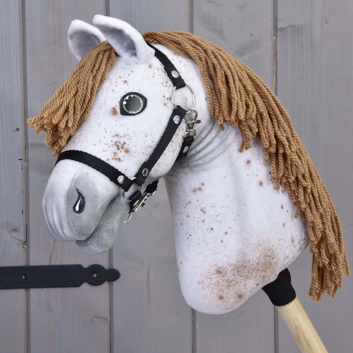Hobby Horse Rufus z czarnym kantarem od Hobby Horse LarDen ma ręcznie wykonaną głowę z białej nakrapianej tkaniny, brązową grzywę z włóczki, oczy z guzików i czarny kantar na drewnianym kiju, na tle szarego drewna.