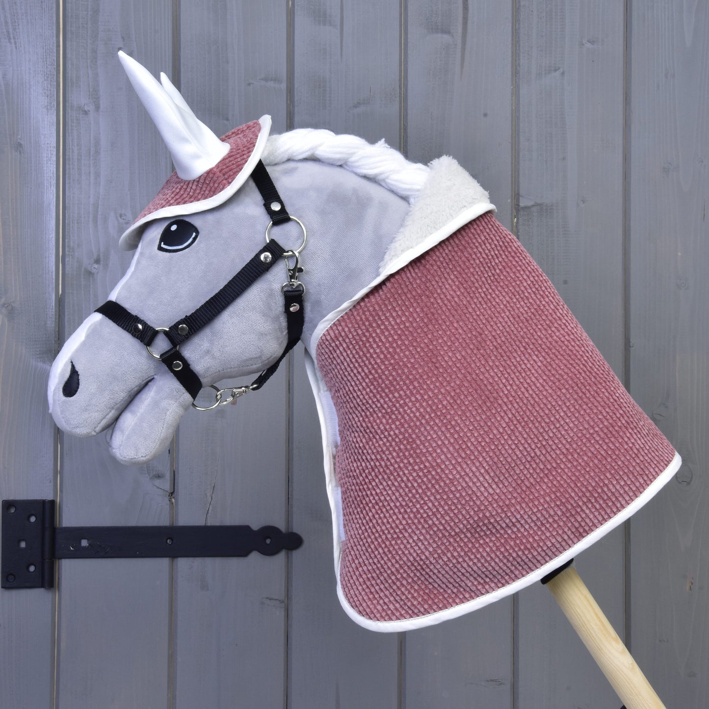 Zestaw Manchester Tea Rose od Hobby Horse LarDen przedstawia pluszowego jednorożca z białym rogiem, czarną uzdą, różowoczerwonym okryciem, nausznikiem i białą grzywą na drewnianym kiju, pokazany na tle szarej drewnianej ściany.