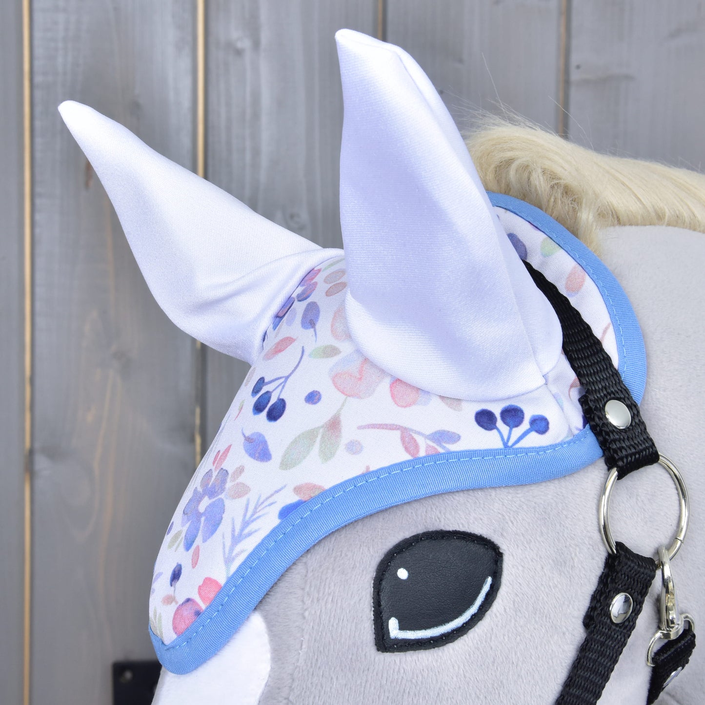 Close-up of the Set Vanessa NEW by Hobby Horse LarDen: a plush hobby horse head with a pastel floral ear bonnet featuring soft white ears, shown against a wooden background.