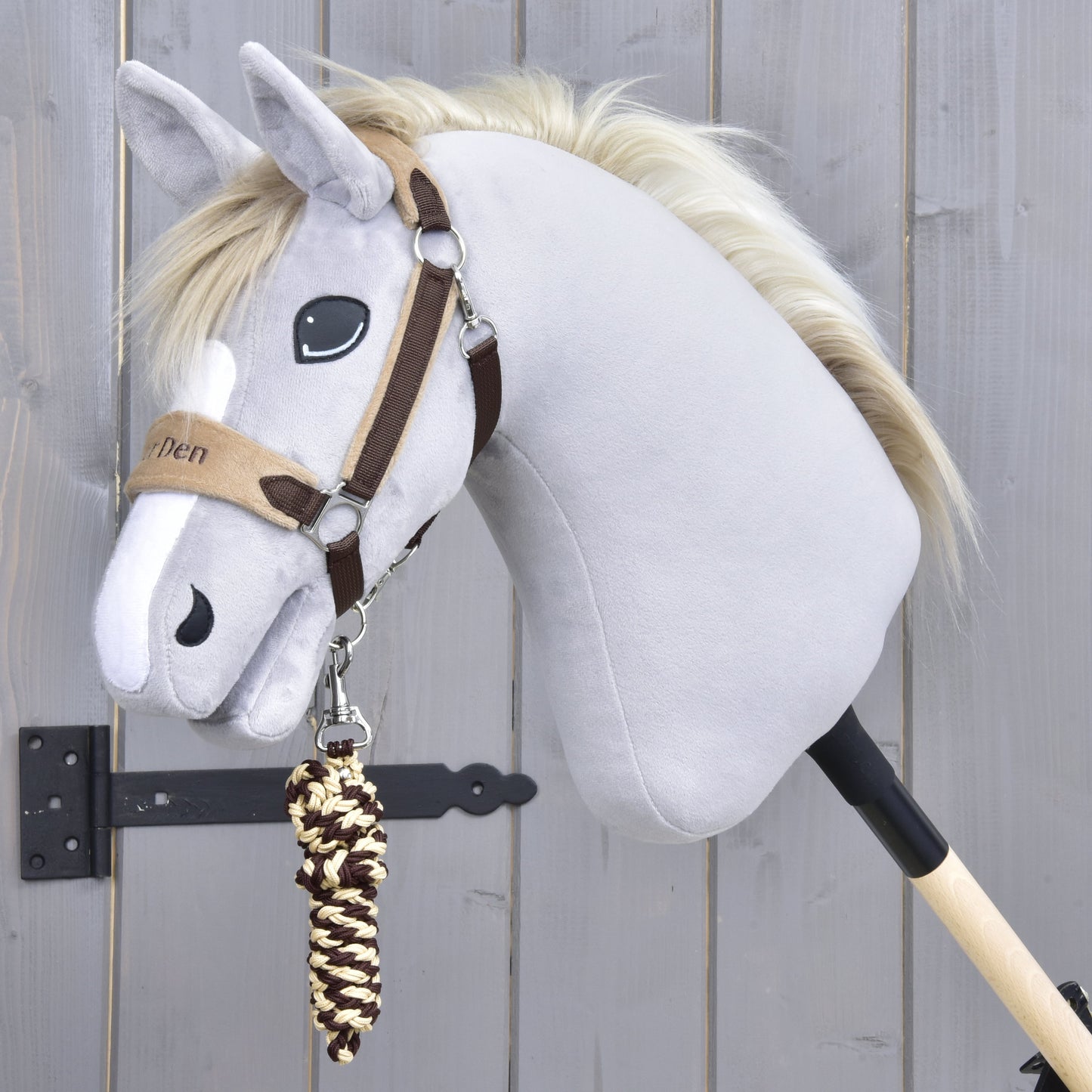 The Set LarDen Exclusive Braun by Hobby Horse LarDen features a gray hobby horse with a bridle, fleece noseband, and cream-brown braided lead rope on a wooden stick, photographed against a light gray wooden wall.