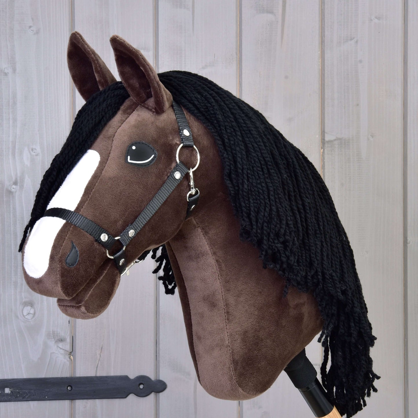 The Hobby Horse LarDen Hobby Horse Sisi with Black Halter, featuring dark brown fur, a white facial stripe, black yarn mane, and resembling a young Hanoverian sport horse, is displayed against a wooden panel background.