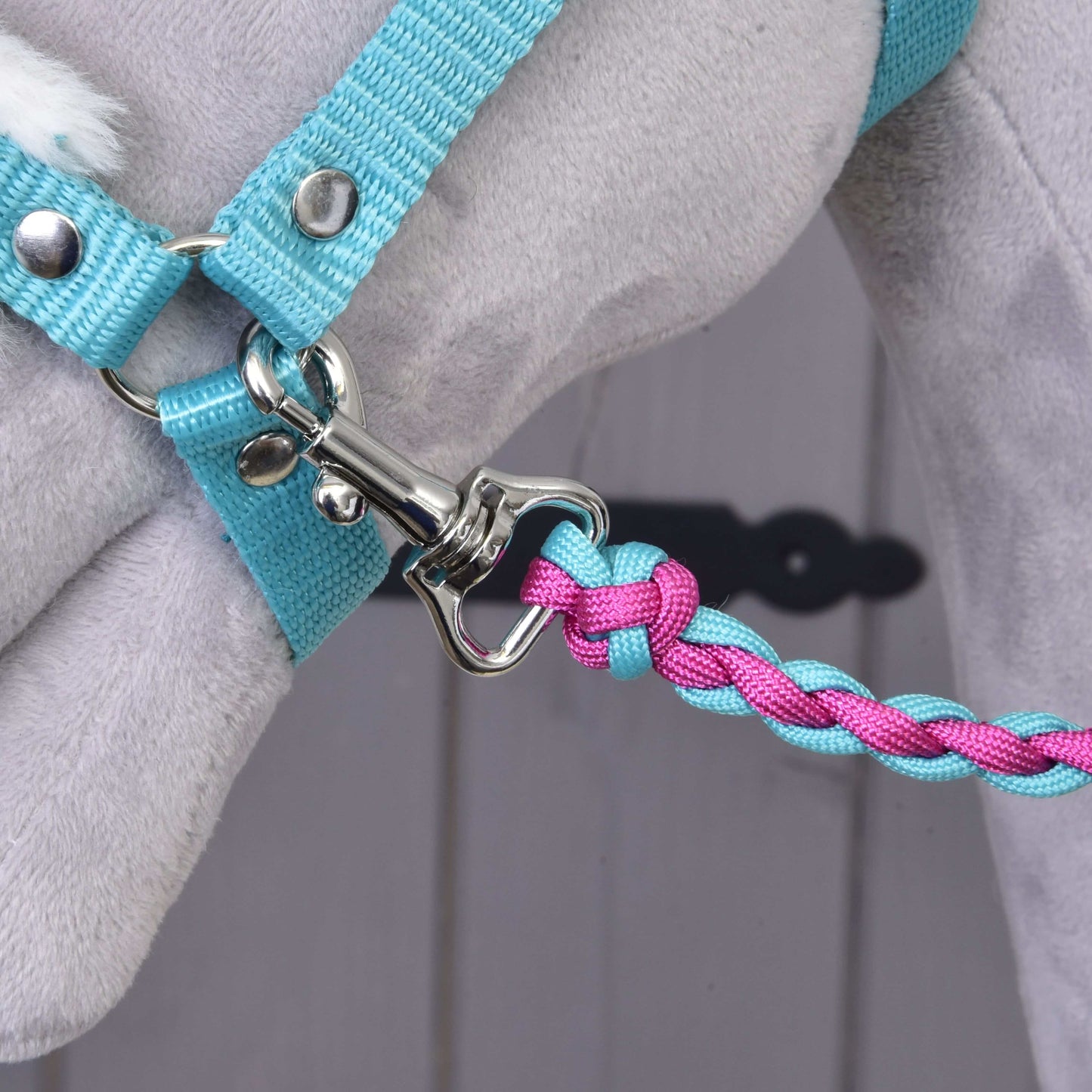Close-up of the Set Perfect Combination Turquoise lead rope by Hobby Horse LarDen, braided in blue and pink and clipped to a light blue harness on a gray plush animal—ideal for hobby horse accessories. Blurred background.