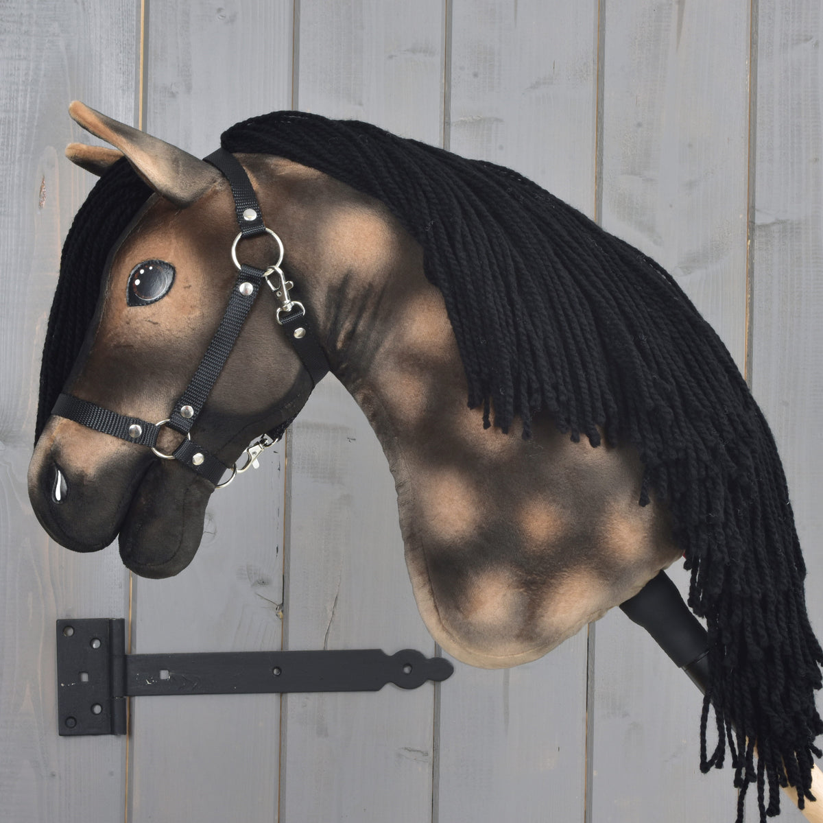 Realistyczny Hobby Horse Stormy z czarnym kantarem od Hobby Horse LarDen, inspirowany ogierem hanowerskim i z czarną włóczkową grzywą, zamocowany do szarej drewnianej ściany na czarnym uchwycie.