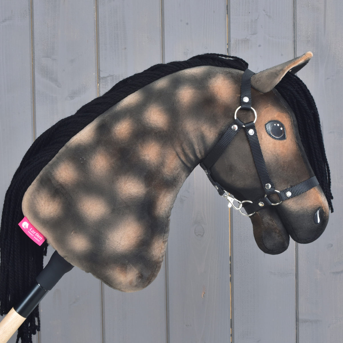 Hobby Horse LarDen „Hobby Horse Stormy z czarnym kantarem” prezentuje cętkowaną brązowo-czarną pluszową głowę inspirowaną ogierem hanowerskim, z czarną włóczkową grzywą i czarnym kantarem, na tle jasnego drewna.