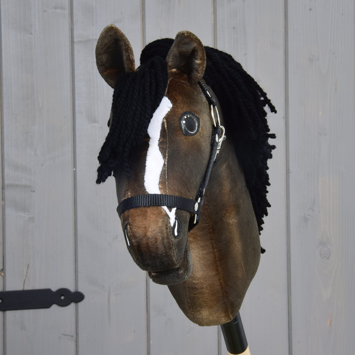 The Hobby Horse LarDen Sundance features a brown coat, white blaze, black yarn mane, and black halter, resembling a spirited Thoroughbred stallion, shown against a light gray wooden plank background.