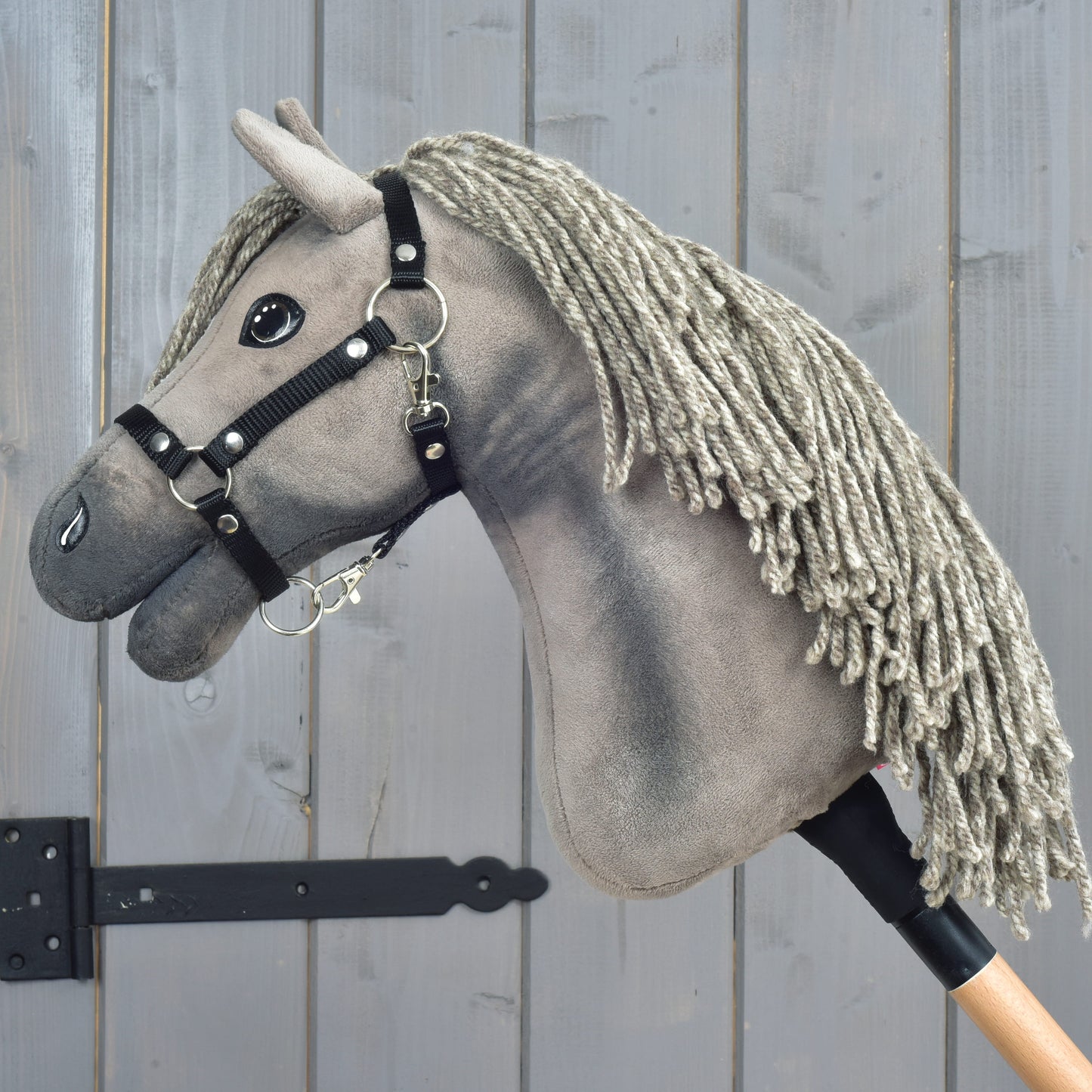 The Hobby Horse LarDen Hobby Horse Swing with black halter, size S, is inspired by the elegance of a Hanoverian stallion and features a gray yarn mane, black bridle, and button eyes. It is mounted on a wooden stick and photographed against a gray wooden background.
