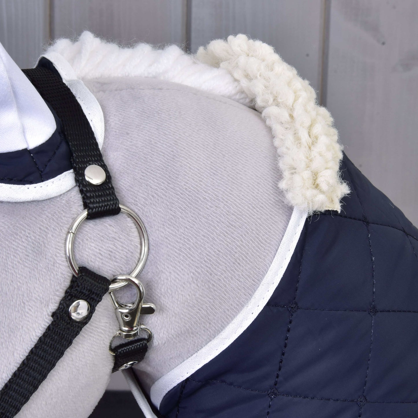 Zbliżenie barku psa w płaszczu Zestaw Termo Niebieski od Hobby Horse LarDen, z pikowanej granatowej tkaniny, z białym wykończeniem i puszystym kołnierzem oraz czarnymi szelkami z metalowym kółkiem. W tle widoczna drewniana boazeria.