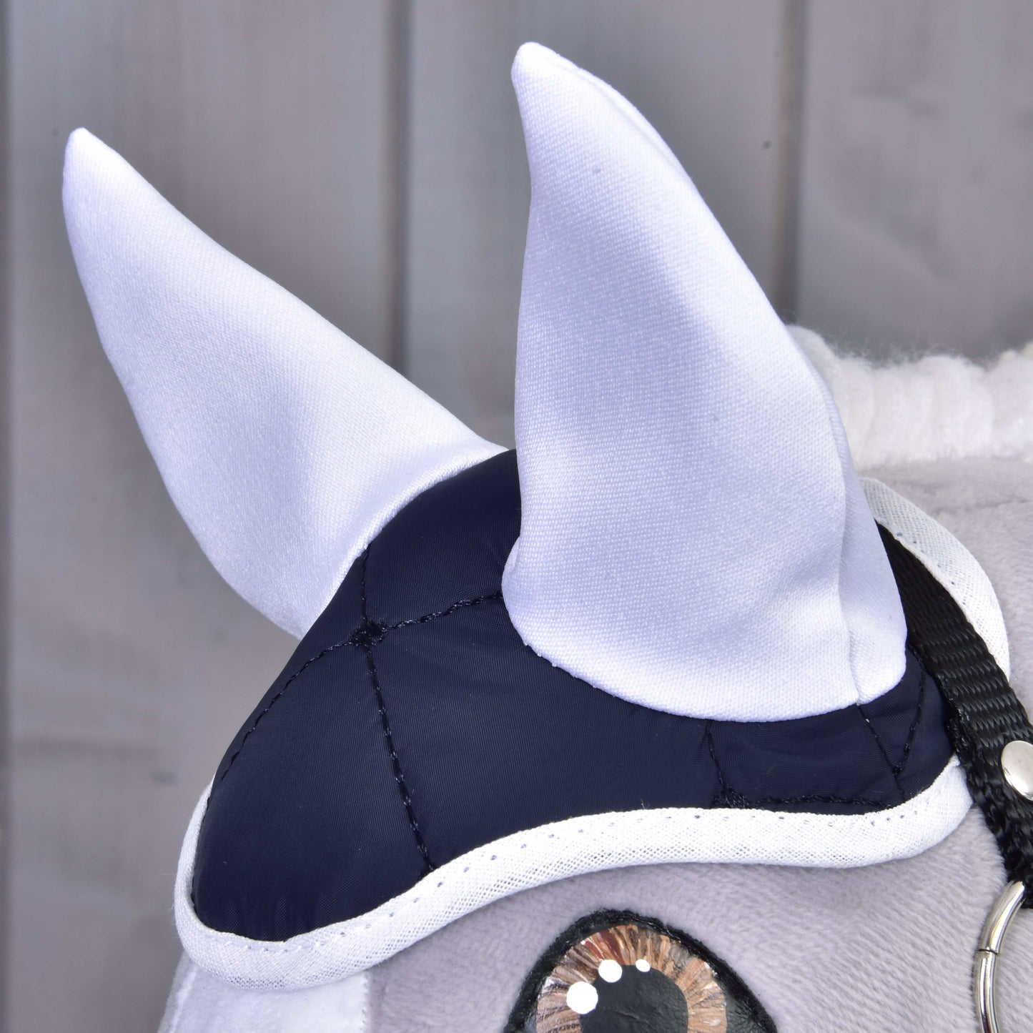 Zbliżenie pluszowej głowy konia w eleganckim Zestawie Termo Niebieskim od Hobby Horse LarDen, z białymi uszami, granatowo-białym czepkiem i czarną uzdą. Idealna dekoracja na tle jasnych drewnianych paneli.