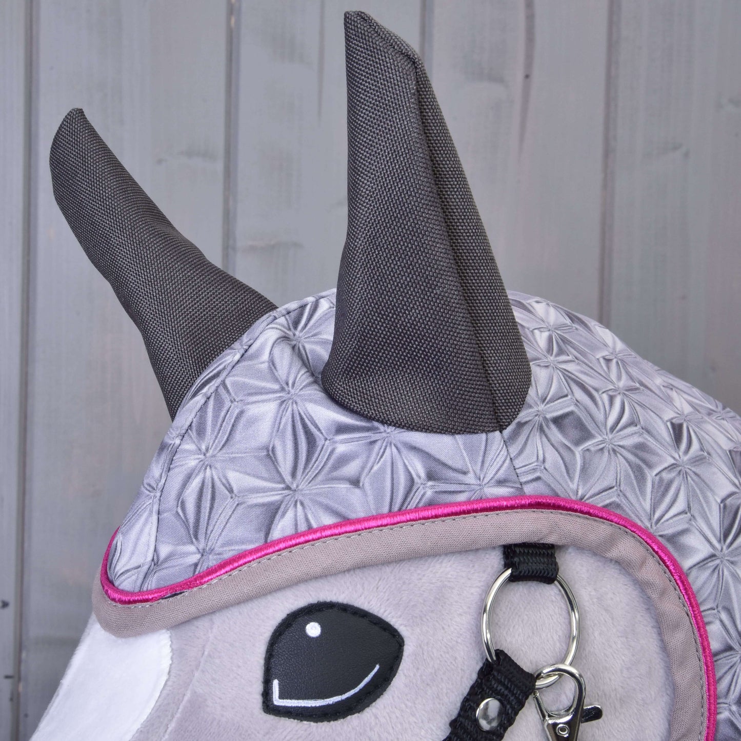 A close-up of a plush horse toy wearing the Hobby Horse LarDen Raincoat Valentine, featuring a patterned gray design with dark gray ears, pink trim, and a black and silver bridle, against a wooden background.