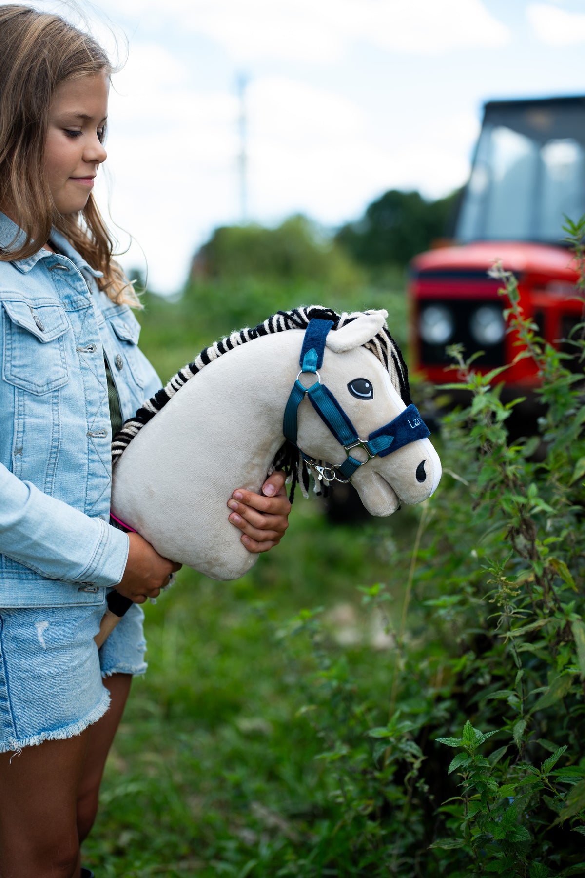 Lifestyle photo: a girl in a denim jacket holds the Hobby Horse Aurora with Black Halter by Hobby Horse LarDen beside a hedge, a red tractor blurred in the background - cream-white plush head, black and white striped yarn mane - shown here with the fur-lined LarDen Blue halter.