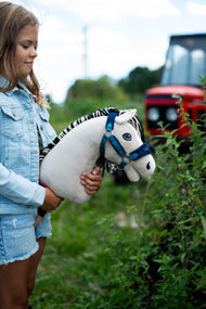Dziewczynka w dżinsowej kurtce trzyma przy żywopłocie Hobby Horse Aurora z czarnym kantarem od Hobby Horse LarDen, z rozmytym czerwonym traktorem w tle – kremowobiała pluszowa głowa, czarno-biała grzywa z włóczki – pokazany z podszytym futerkiem kantarem LarDen Blue