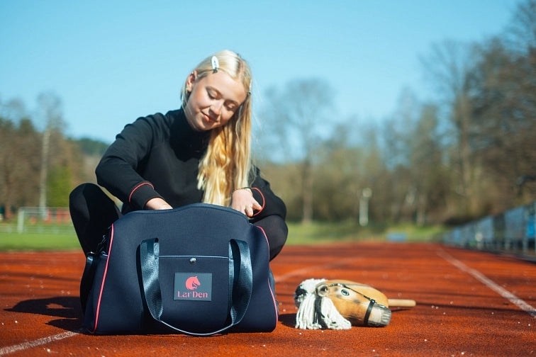 Młoda kobieta z długimi blond włosami siedzi na bieżni i zagląda do granatowej torby sportowej z napisem LarDen. Obok niej na ziemi leży hobby horse, sugerując jej pasję do Hobby Horsing. W tle widoczne są drzewa i linie bieżni.