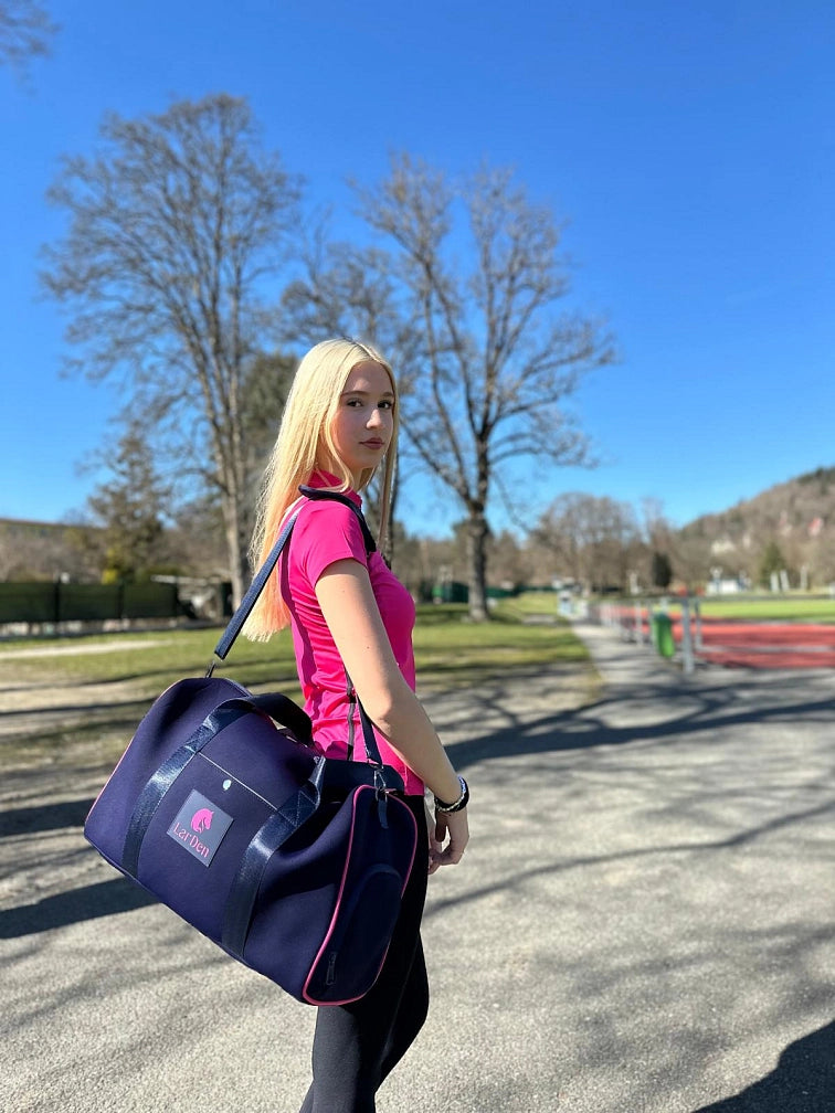 Młoda kobieta z długimi blond włosami, w różowej koszulce sportowej i z granatową torbą sportową, stoi na zewnątrz w słoneczny dzień ze swoim Hobby Horse, gotowa na pełen zabawy dzień Hobby Horsing pod czystym błękitnym niebem i wśród zielonych drzew.