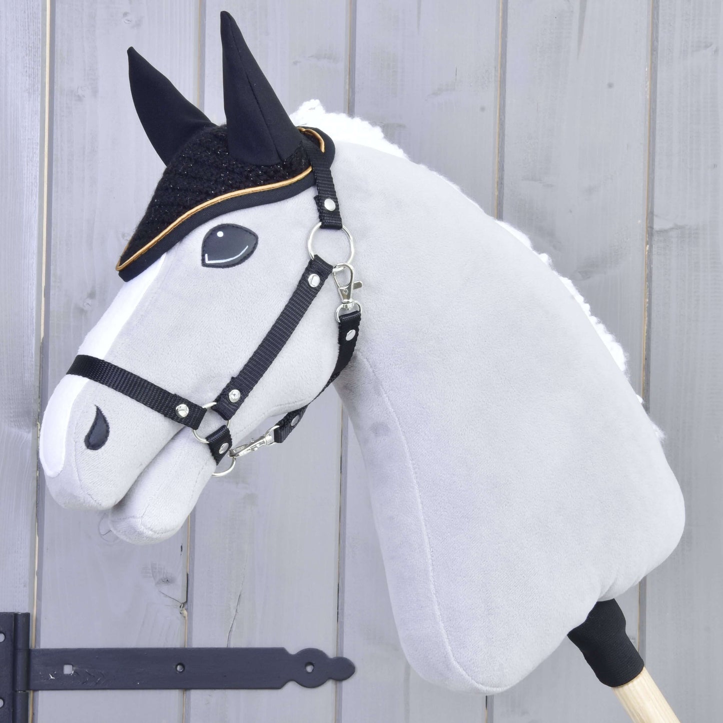 A gray Hobby Horse LarDen with black reins and a white mane wears the Ear bonnet crocheted Black Gold for insect protection, featuring black facial details, set against a light wooden background.