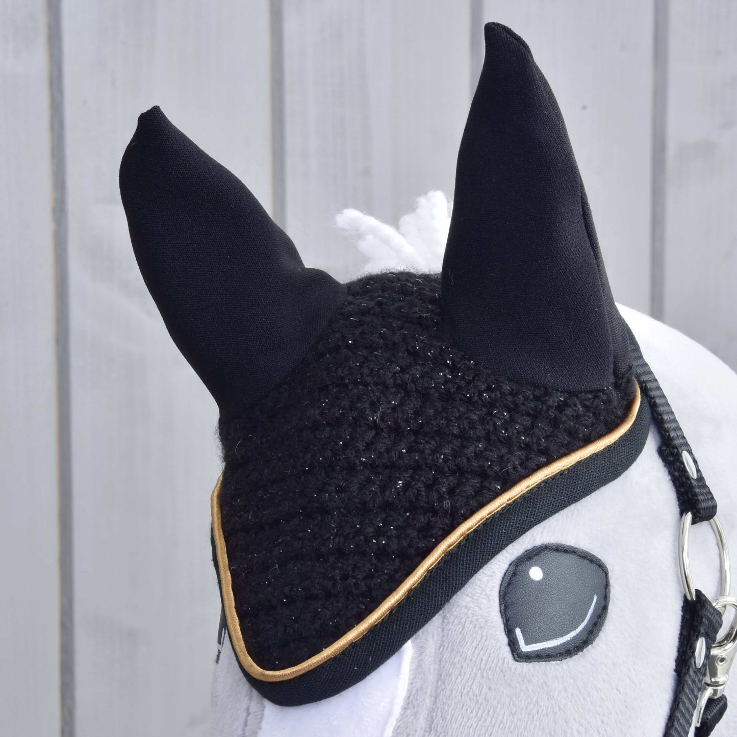 A close-up of a plush horse toy from Hobby Horse LarDen wearing the Ear bonnet crocheted Black Gold for insect protection. The horse features a bridle, white mane, and is set against a light wooden wall.
