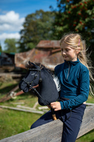 Blond dziewczynka w turkusowej bluzce siedzi na drewnianej poręczy w słonecznym gospodarstwie, trzymając Hobby Horse Bond z czarnym kantarem od Hobby Horse LarDen – czarna pluszowa głowa, ciemnobrązowa grzywa z włóczki – pokazany z ozdobnym ogłowiem bezwędzidłowym