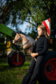 Dziewczynka w czarnym golfie stoi przy zielonych maszynach rolniczych, trzymając Hobby Horse Bren z czarnym kantarem od Hobby Horse LarDen – piaskowobrązowa pluszowa głowa, kremowa grzywa z włóczki – pokazany z napierśnikiem Espresso z futerkiem