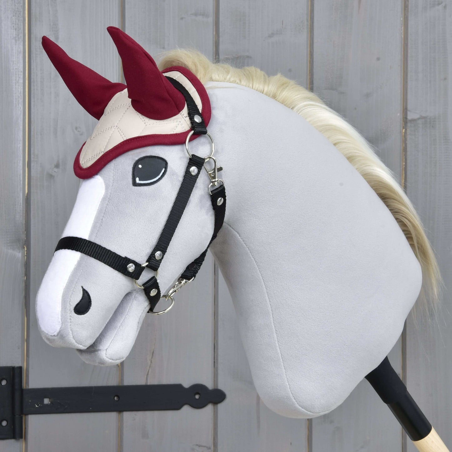 Pluszowy Hobby Horse od Hobby Horse LarDen z białym tułowiem, czarną uzdą i kremową grzywą ma na sobie Nauszniki Bordowa Gwiazda do ochrony przed owadami, pokazany na kiju na drewnianym tle.