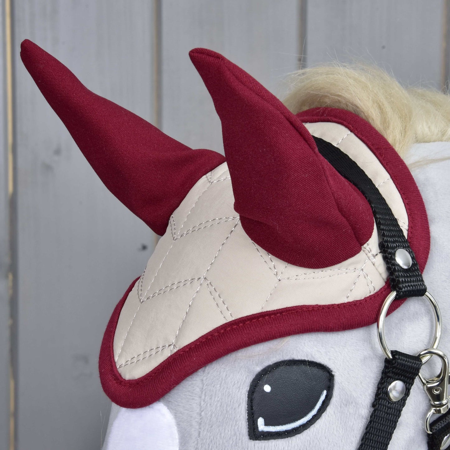 Zbliżenie Hobby Horse LarDen z Nausznikami Bordowa Gwiazda, z beżowym pikowaniem do ochrony przed owadami, dwoma stojącymi czerwonymi kształtami przypominającymi rogi i czarną uzdą. W tle jasne drewniane panele.