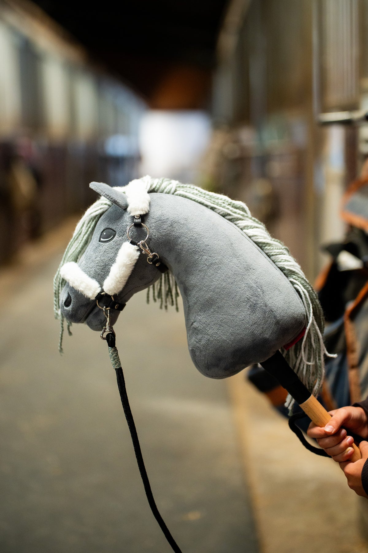 Detail photo: the head of the Hobby Horse Calypso with Black Halter by Hobby Horse LarDen held up in a stable aisle, the stalls blurred behind - grey plush, pale grey yarn mane, dark eye - shown here with the fur-lined Teddy Classic halter and the black lead rope.
