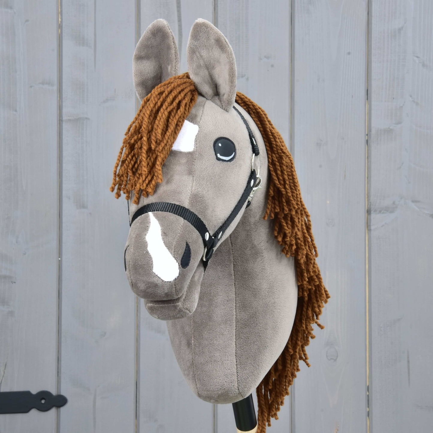 The Hobby Horse LarDen Hobby Horse Chanell with Black Halter features a gray coat, brown yarn mane, white face patches, large expressive eyes, and is displayed against a gray wooden background.