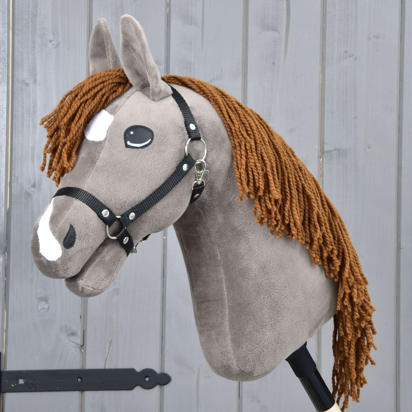 The Hobby Horse LarDen Chanell features a soft plush build, a black halter, white facial markings, brown yarn mane, and cartoon eyes, shown against a light gray wooden wall.