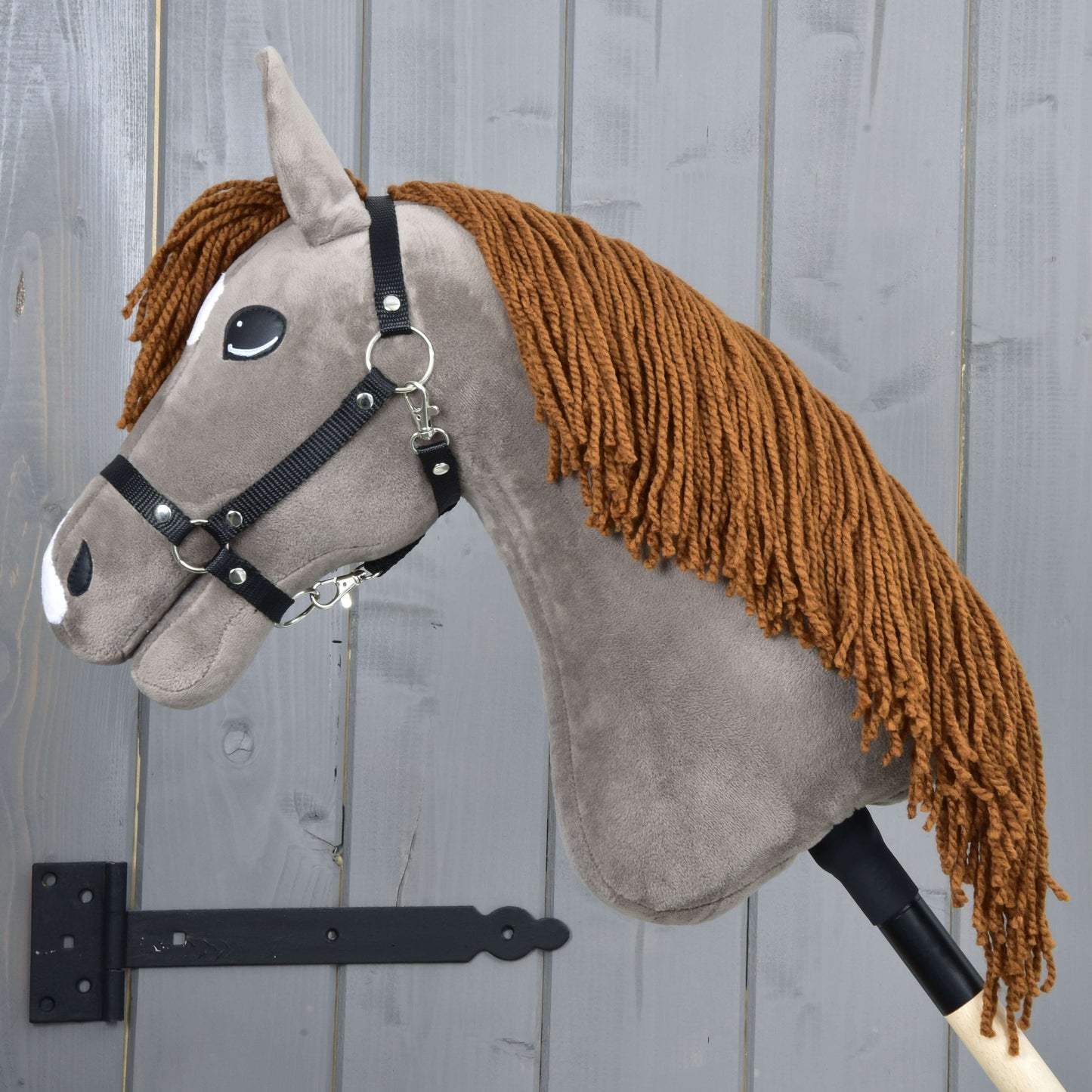 The Hobby Horse LarDen Chanell with Black Halter features a gray fabric head, brown yarn mane, black halter, white felt accents, and is mounted on a wooden stick in front of a gray wooden plank wall.