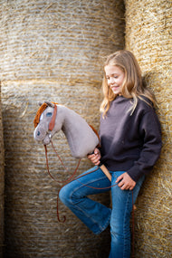 Uśmiechnięta dziewczynka w ciemnobrązowej bluzie opiera się o okrągłe bele słomy, trzymając Hobby Horse Chanell z czarnym kantarem od Hobby Horse LarDen – szara pluszowa głowa, niebieskie oko, imbirowa grzywa z włóczki – pokazany z kompletnym westernowym ogłowiem z jednym nausznikiem
