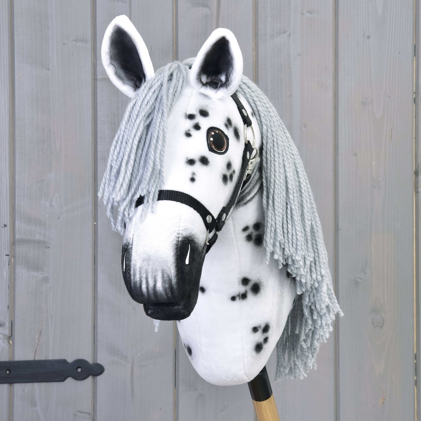 The Hobby Horse Chantal with Black Halter by Hobby Horse LarDen is a handcrafted mare inspired by the Knabstrupper breed, featuring a white face, black spots, long gray mane, and mounted on a wooden stick in front of a gray wall.