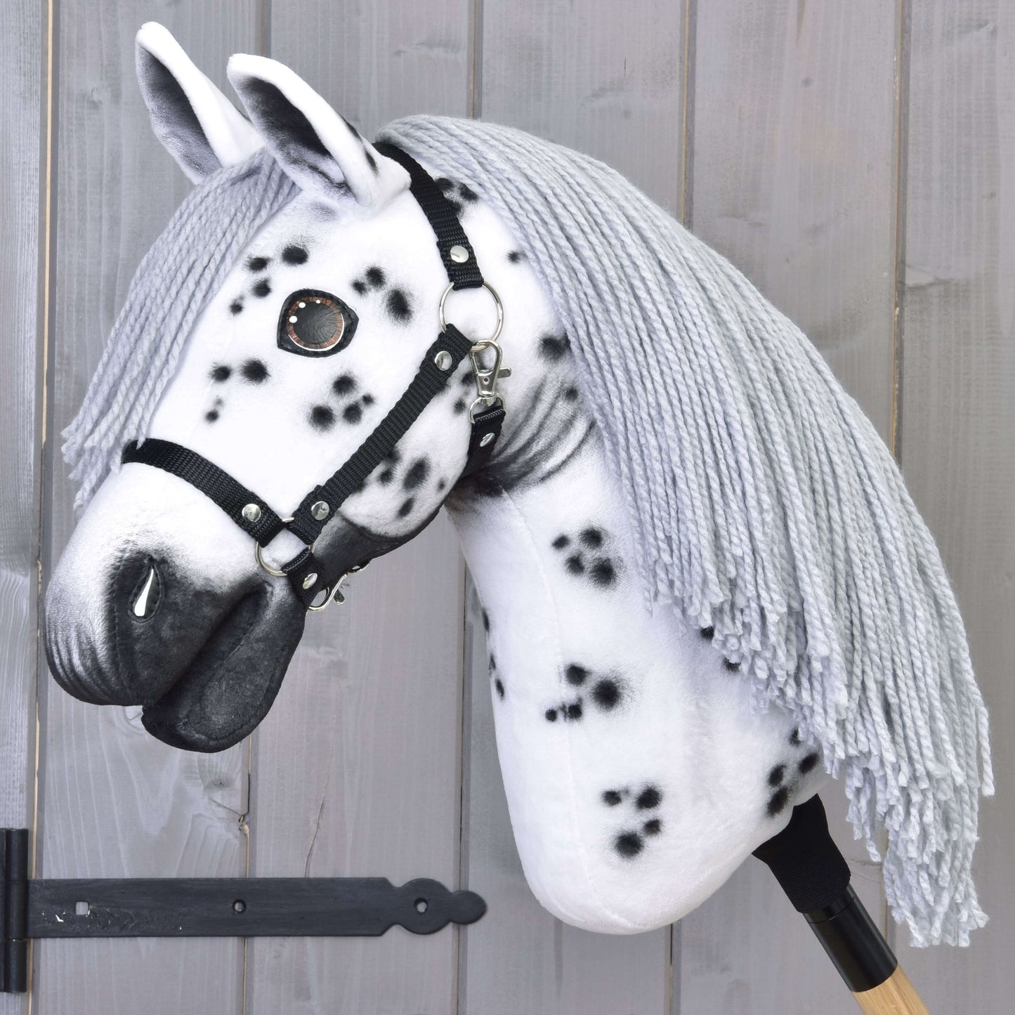 The Hobby Horse LarDen "Chantal with Black Halter" showcases a handcrafted white Knabstrupper-style head with black spots, gray yarn mane, black halter, and wooden stick handle, set against a light wooden plank background.
