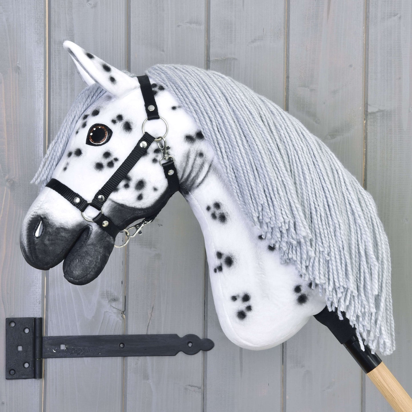 The Hobby Horse LarDen "Chantal" features white fur with black spots, a gray yarn mane, and comes with a black halter. This handmade Knabstrupper hobby horse is mounted on a wooden stick and posed against a gray wooden wall.