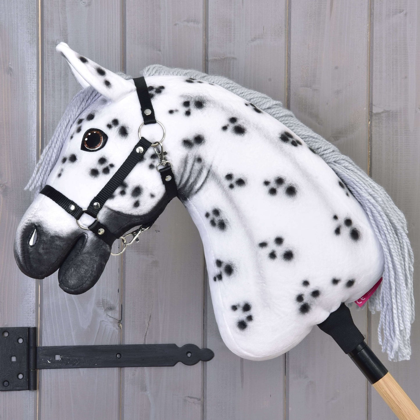 The Hobby Horse LarDen 'Hobby Horse Chantal with Black Halter' features a white coat with black spots, gray yarn mane, black halter, and is mounted on a wooden stick against a light gray wooden wall.