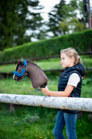 Dziewczynka w granatowej pikowanej kamizelce stoi za białą poręczą padoku, trzymając Hobby Horse Chianti z czarnym kantarem od Hobby Horse LarDen, z przystrzyżonym zielonym żywopłotem w tle – ciemnobrązowa pluszowa głowa, ciemna grzywa z włóczki – pokazany z podszytym futerkiem kantarem Teddy Petrol
