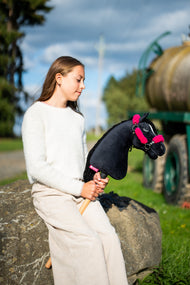 Dziewczynka w kremowym swetrze siedzi na głazie przy polnej ścieżce, trzymając Hobby Horse Cloony z czarnym kantarem od Hobby Horse LarDen – czarna pluszowa głowa, biała gwiazdka, czarna grzywa z włóczki – pokazany z czarnym kantarem ze skóry ekologicznej z pełnym różowym futerkiem