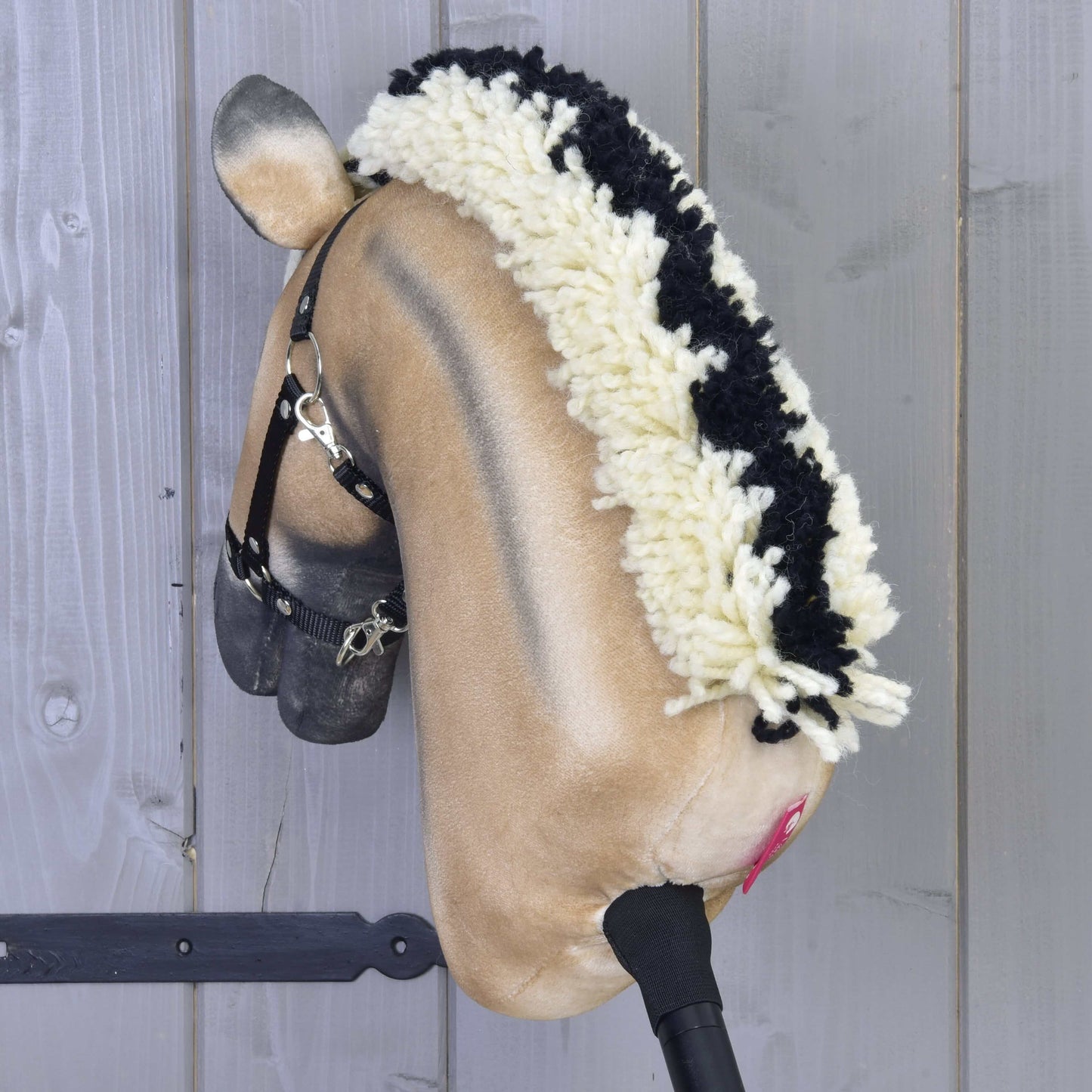 Hobby Horse LarDen „Colby” ma beżowy pyszczek, czarno-białą włóczkową grzywę, czarny kantar i różowe nozdrza, osadzony na drewnianym kiju na tle szarej drewnianej ściany.