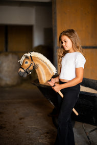 Dziewczynka w białej bluzce stoi w stajni, trzymając Hobby Horse Colby z czarnym kantarem od Hobby Horse LarDen – palomino pluszowa głowa, gęsta kremowa grzywa z włóczki, czarny kantar ze srebrnymi okuciami