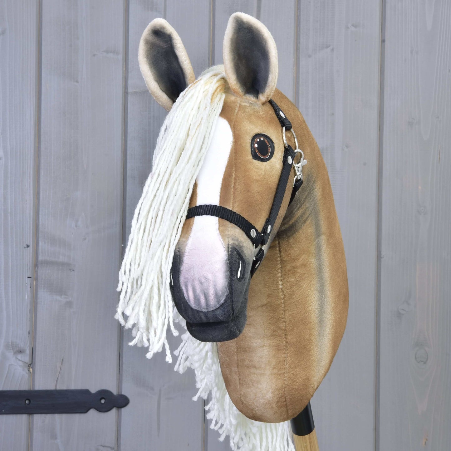 Hobby Horse Connie z Czarnym Kantarem od Hobby Horse LarDen ma ręcznie wykonany jasnobrązowo-biały pysk, duże oczy, długą białą grzywę z włóczki i czarny kantar, zamocowany na kiju przed szarą drewnianą ścianą.