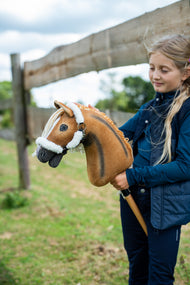 Dziewczynka w granatowej kamizelce stoi przy drewnianym ogrodzeniu padoku, trzymając Hobby Horse Connie z czarnym kantarem od Hobby Horse LarDen – kasztanowata pluszowa głowa z ciemną pręgą grzbietową, kremowa grzywa z włóczki – pokazany z podszytym futerkiem kantarem Teddy Classic