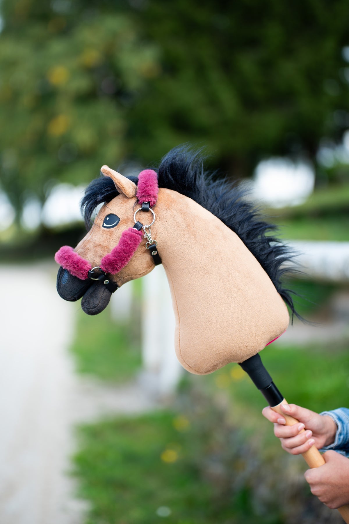 Głowa Hobby Horse Cookie z czarnym kantarem od Hobby Horse LarDen uniesiona przy ścieżce wśród drzew – piaskowy plusz, długa czarna grzywa z włóczki – pokazana z podszytym futerkiem kantarem Teddy Maroon