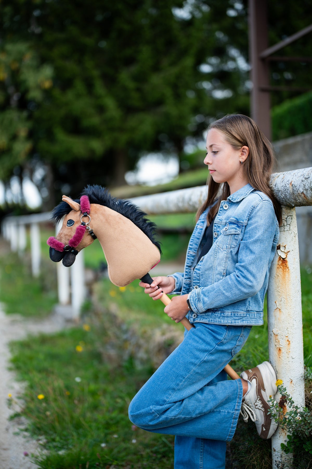 Dziewczynka w dżinsowym stroju opiera się o białą poręcz padoku, trzymając Hobby Horse Cookie z czarnym kantarem od Hobby Horse LarDen – piaskowa pluszowa głowa, czarna grzywa z włóczki – pokazany z podszytym futerkiem kantarem Teddy Maroon