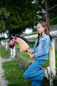 Dziewczynka w dżinsowym stroju opiera się o białą poręcz padoku, trzymając Hobby Horse Cookie z czarnym kantarem od Hobby Horse LarDen – piaskowa pluszowa głowa, czarna grzywa z włóczki – pokazany z podszytym futerkiem kantarem Teddy Maroon