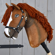 Introducing the Hobby Horse Cooper with Black Halter from Hobby Horse LarDen—a brown hobby horse inspired by an Oldenburg sport horse. This charming toy features a white blaze on its face, thick dark brown yarn hair, and comes with a black nylon halter accented by metal hardware. Attached to a wooden stick and pictured against grey wooden planks, it's perfect for imaginative dressage play.
